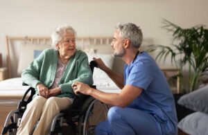 Elderly residents seated in a common room, engaged in discussion with staff members at a long table.